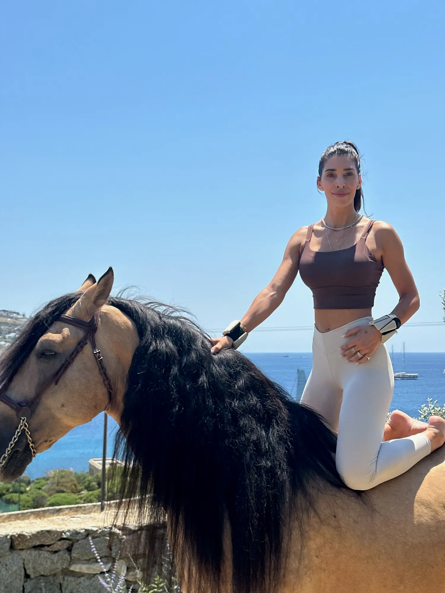 Demy sitting cross-legged on a horse by the sea in Mykonos