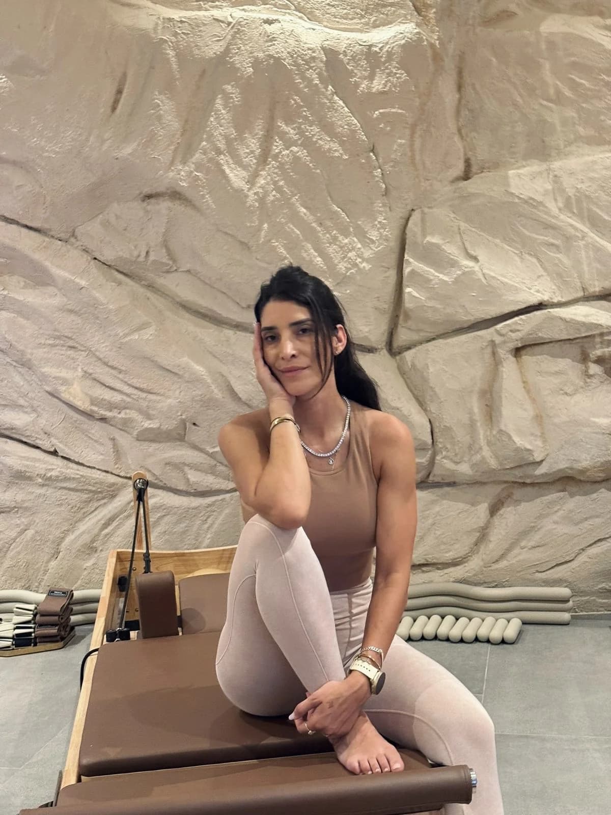 Demy seated on a wooden reformer against the sculpted stone wall of the CÉLINE studio, in warm beige tones